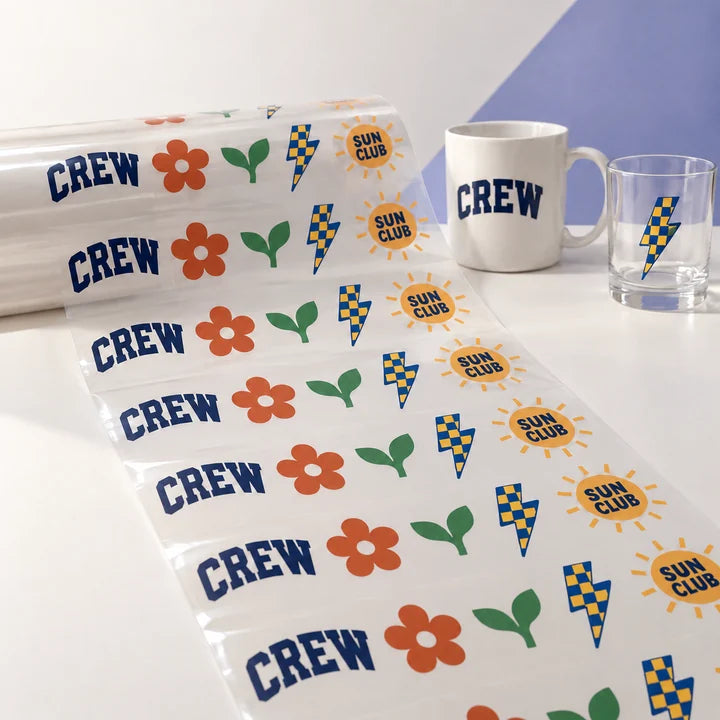 Mockup: long printed UV-DTF decal sheet unrolling across a table, with a mug and glass as examples